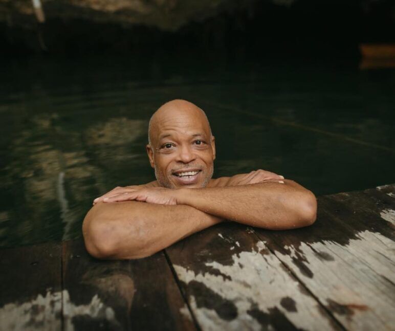 An older black man lounges in the water on the edge of a dock
