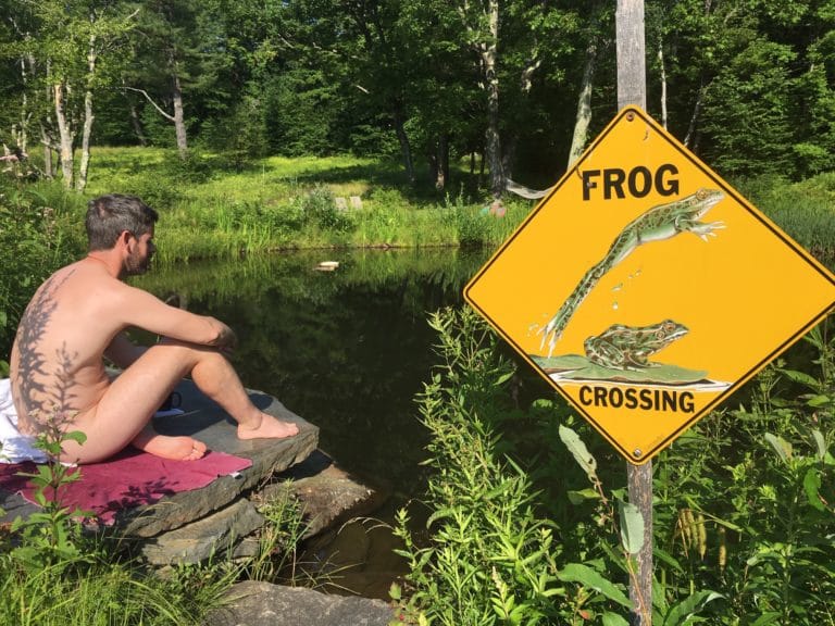 A shirtless man sits next to a pond. A sign near him says "Frog crossing"