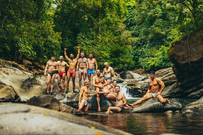 A group of men in swimsuits stand on the edge of a river in the jungle