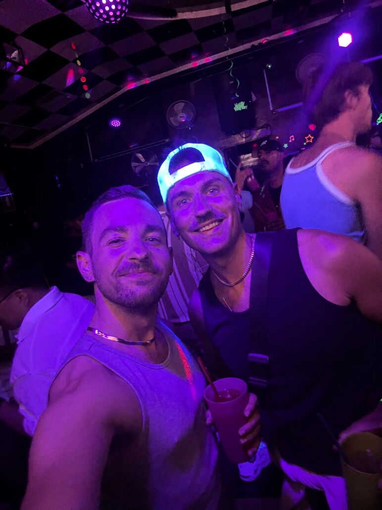 Ian and their partner, Smith, take a selfie under blacklight at a crowded gay bar. They’re smiling with drinks in hand, surrounded by other partygoers under purple and neon lighting.