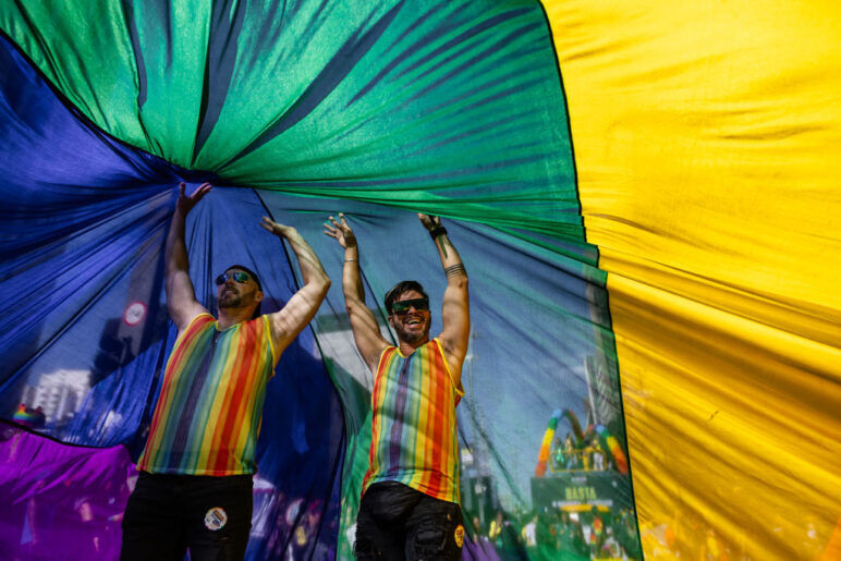Two men wearing rainbow tshirts stand under a giant Pride flag