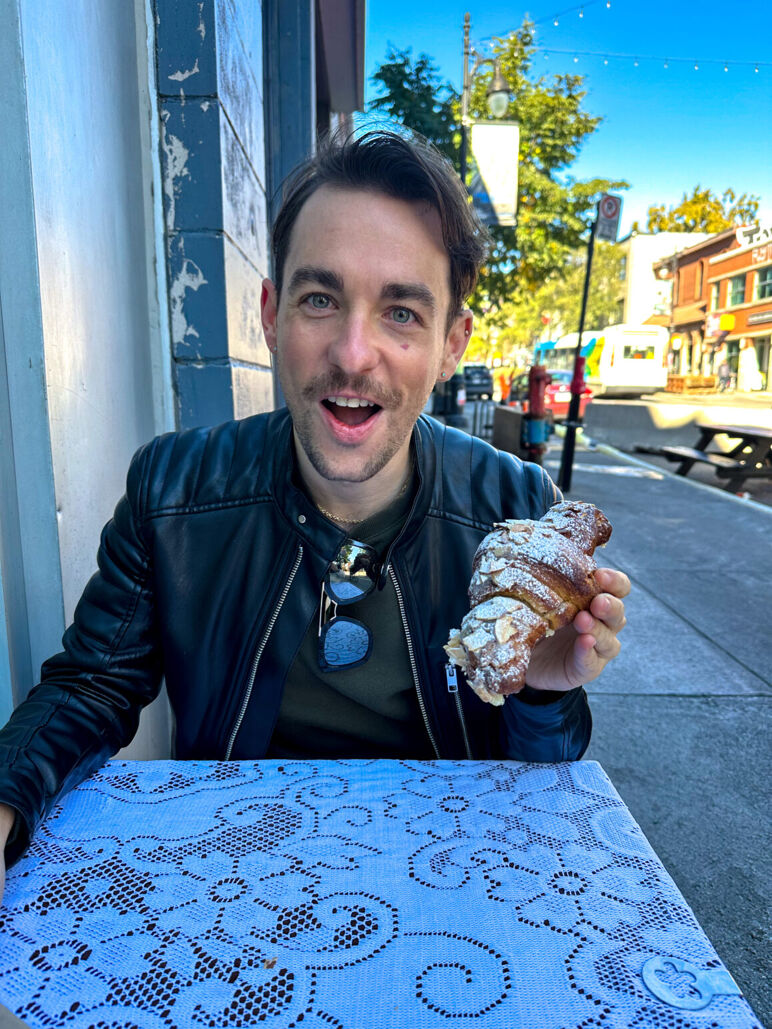 Ian sits at an outdoor cafe table covered with a lace-patterned cloth, holding an almond croissant and smiling with surprise. He wears a black leather jacket and sunglasses, with a sunny street and shops in the background.