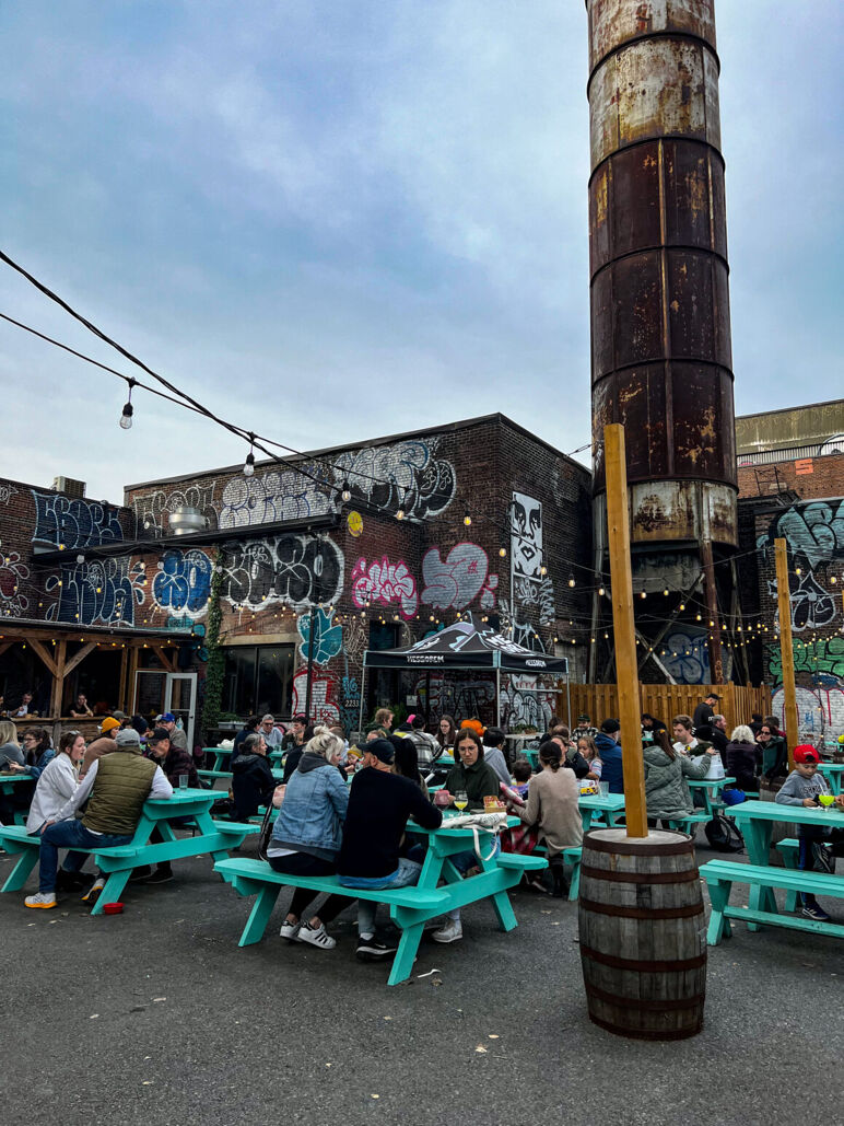 People sit at turquoise picnic tables in a graffiti-covered outdoor beer garden, with string lights and a tall rusty chimney tower in the background. The vibe is casual and artsy.