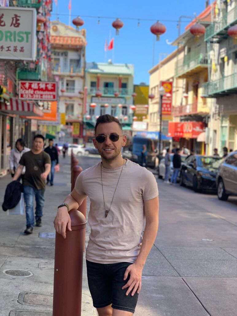 Ian poses on a sunny day in San Francisco’s Chinatown, surrounded by colorful signs, lanterns hanging overhead, and bustling foot traffic.