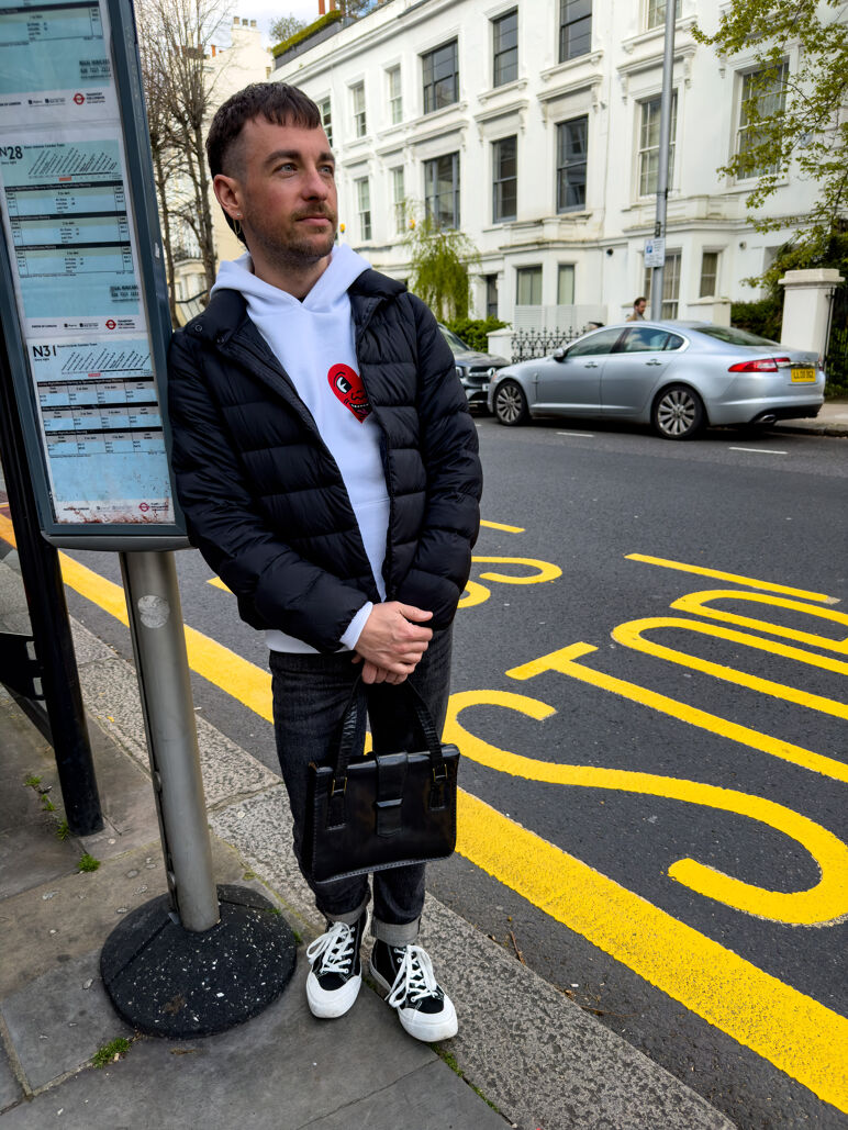 Ian stands at a London bus stop wearing a white hoodie, black puffer jacket, cuffed jeans, and platform sneakers, holding a black handbag. Yellow “BUS STOP” lettering is painted on the road beside him, with white townhouses and parked cars in the background.