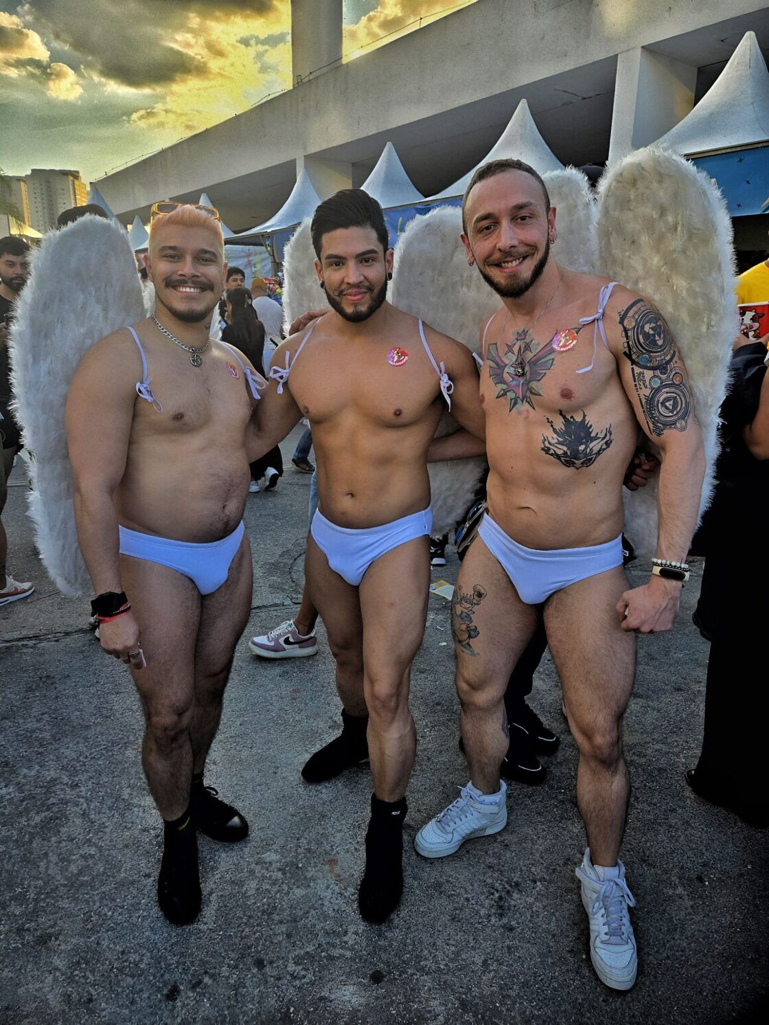 A trio of three muscular and shirtless men smile wearing angel wings and blue speedos.