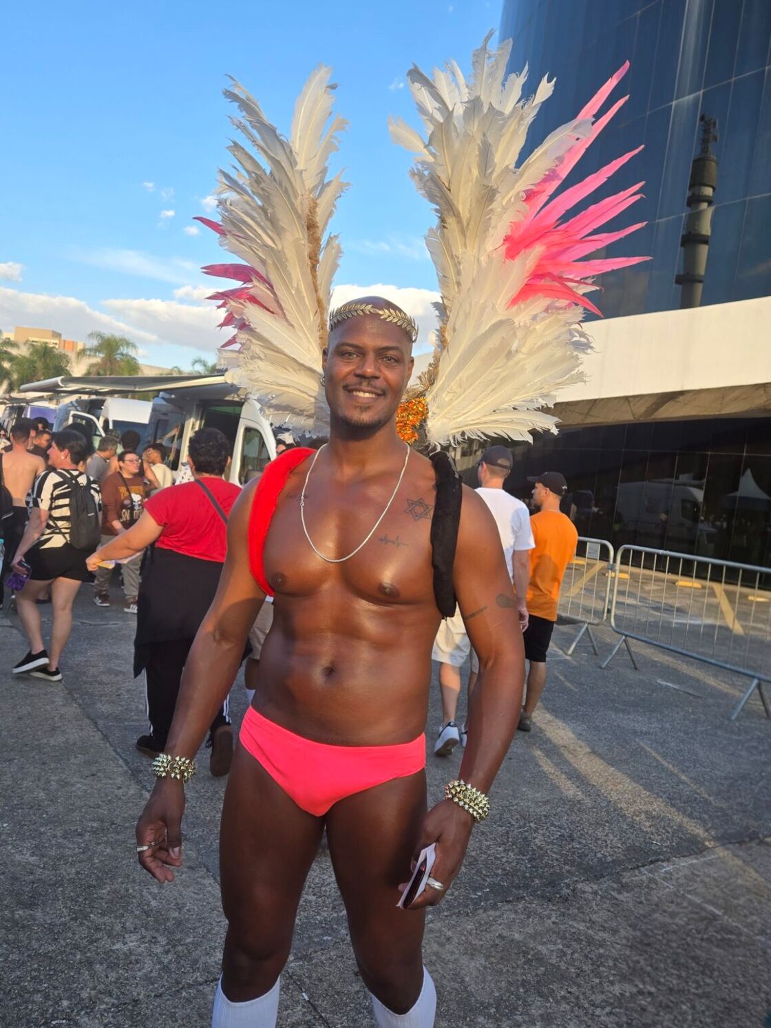A man smiles shirltess, wearing a pink speedo, a golden crown, and white wings.
