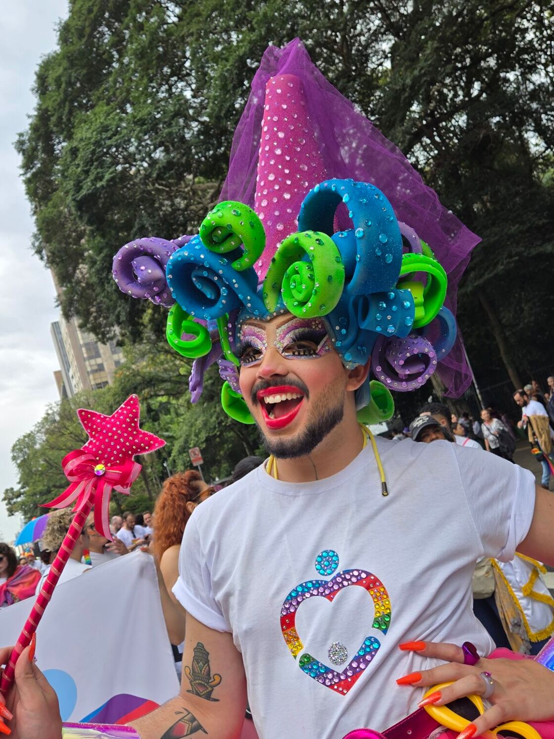 A man smiles wearing drag makeup and a wig of purple, green, and blue swirls, holding a magic wand.