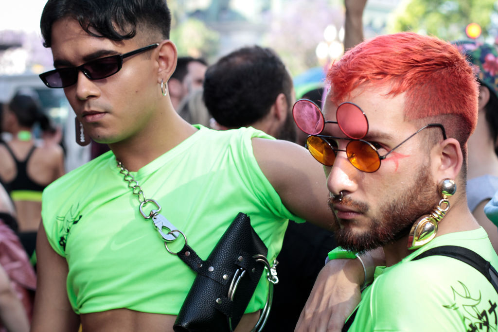 Two Latino men pose at a Pride parade. One is looking at the camera.