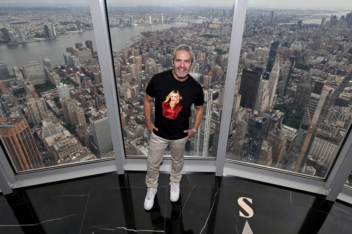Andy Cohen poses with his hands in his pockets in front of the Empire State Building lookout windows.
