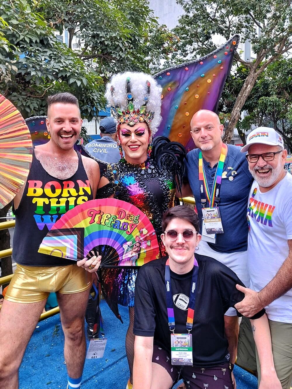 A drag queen with a "Drag Tiffany" fan poses smiling with a group of São Paulo Pride attendees decked in rainbow clothes outdoors.