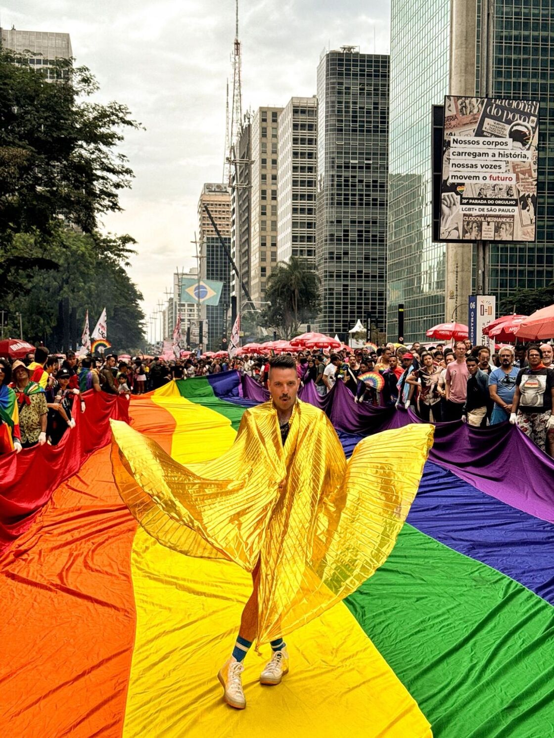 A man dances in a shiny golden sheet while walking a runaway resembling a giant Pride flag in the middle of the São Paulo parade.