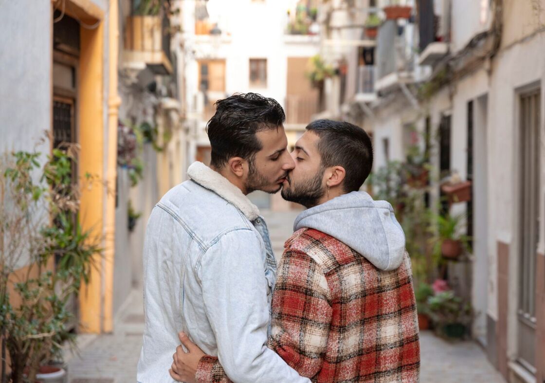 Two men kiss in a sunny corridor near plants and vibrant architecture.