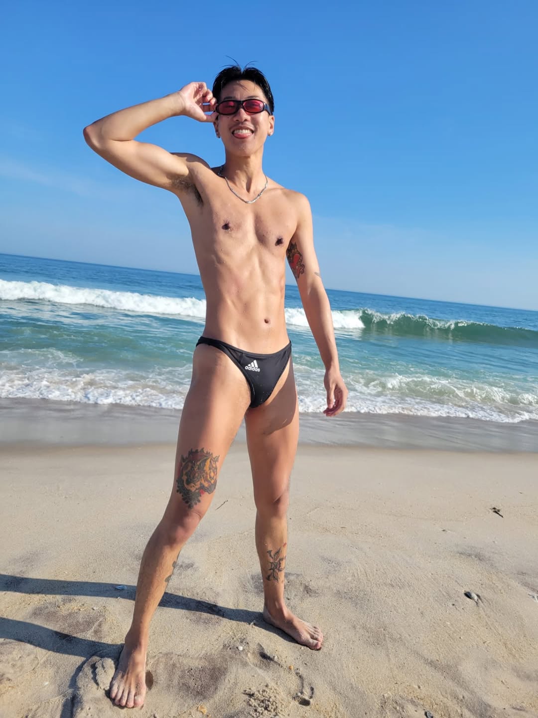 Comedian James Tom poses on the Fire Island beach in a speedo and black sunglasses.