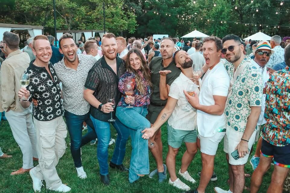 A group of attendees pose together with wine glasses outdoors at Gay Wine Weekend.