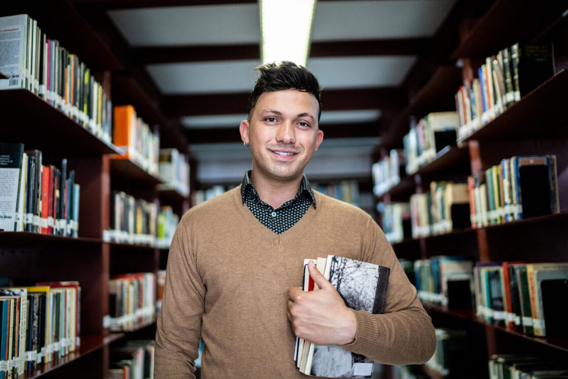 A young man stands smiling in a collared shirt in the middle of a bookstore.