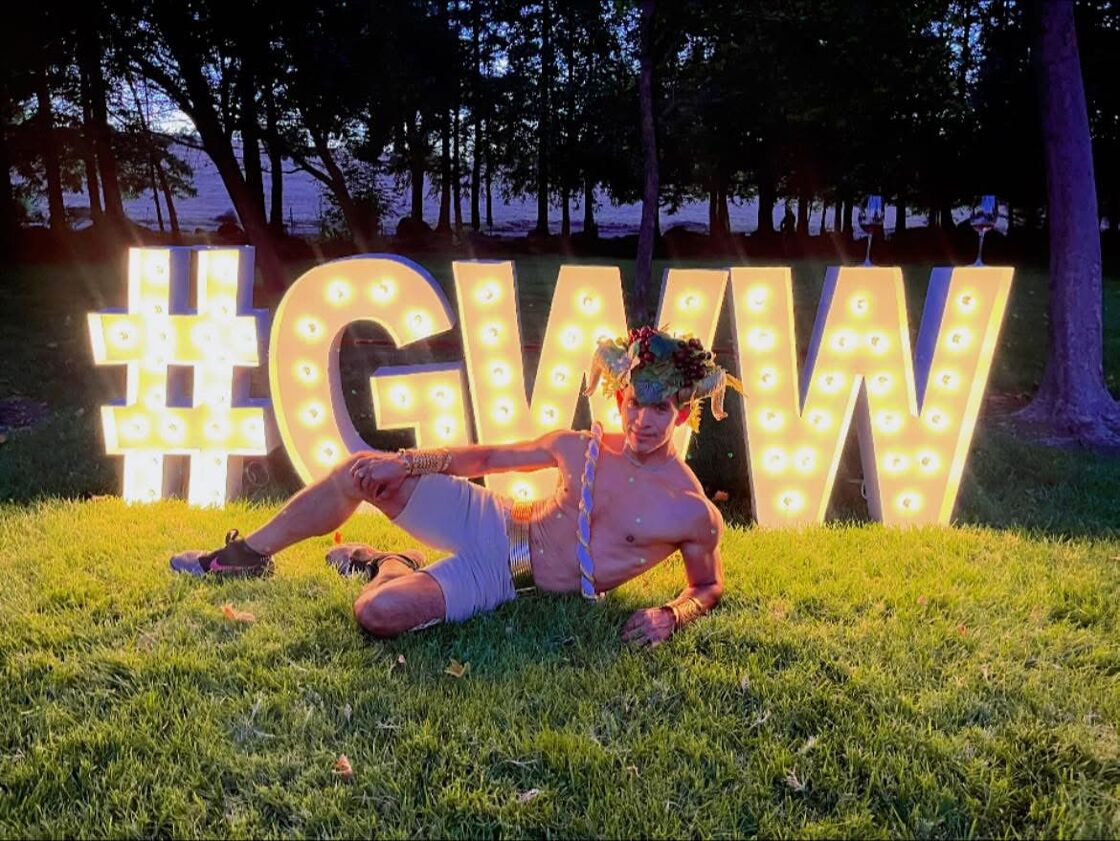 A shirtless man dressed like Dionysus poses in front of a GWW lit up sign. 