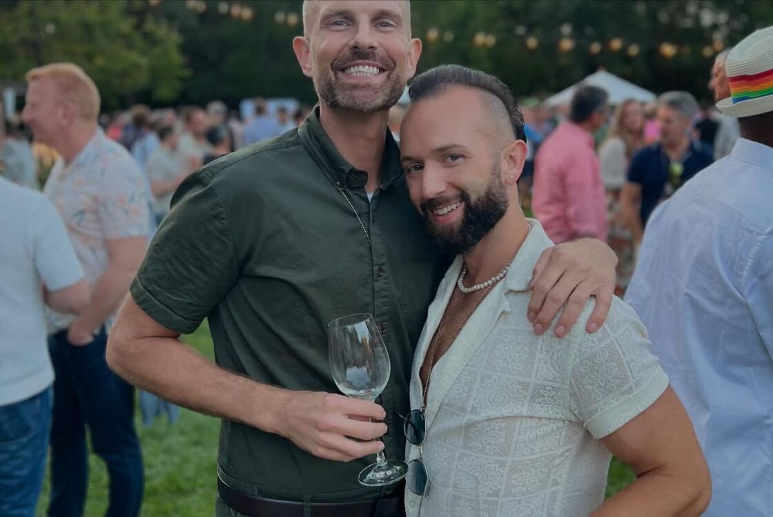 Two men embrace outdoors at Gay Wine Weekend, holding wine glasses.