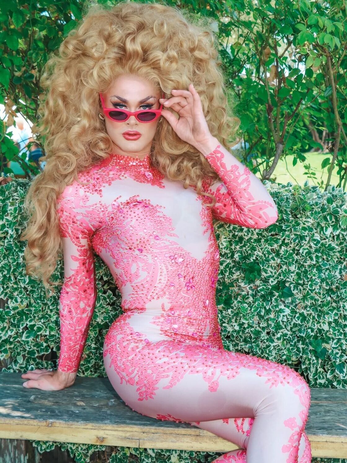 Scarlet Envy (in drag) poses in a pink outfit alongside a shrub at Gay Wine Weekend.