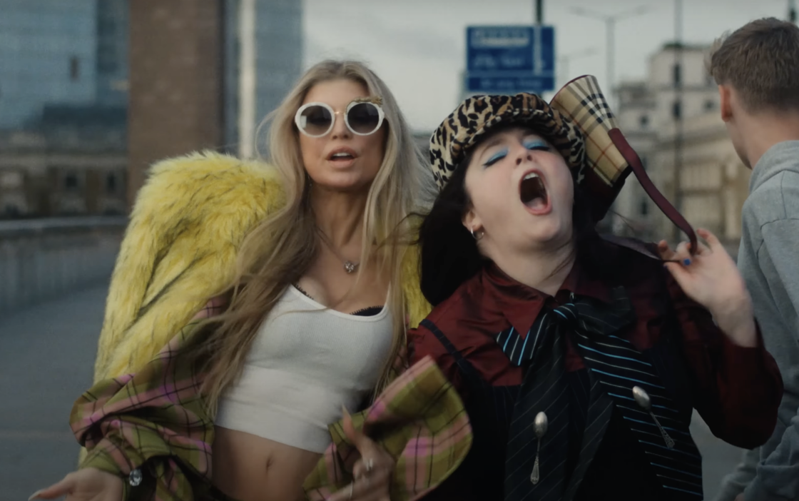 In a screenshot from their new "London Bridge" music video, Fergie and Megan Stalter walk down the London Bridge in fashionable outfits, while a pedestrian bumps into Meg, causing her to fall. 