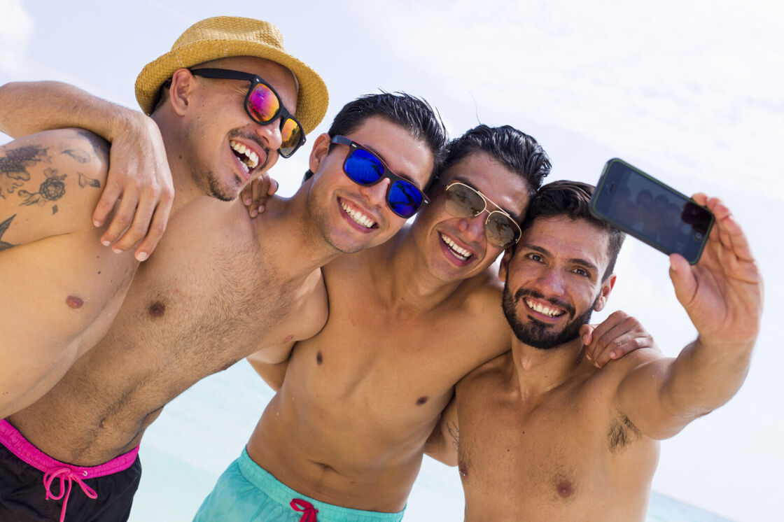 A group of four shirtless men on the beach, wearing sunglasses, stand with their arms around each other and smile for a selfie.