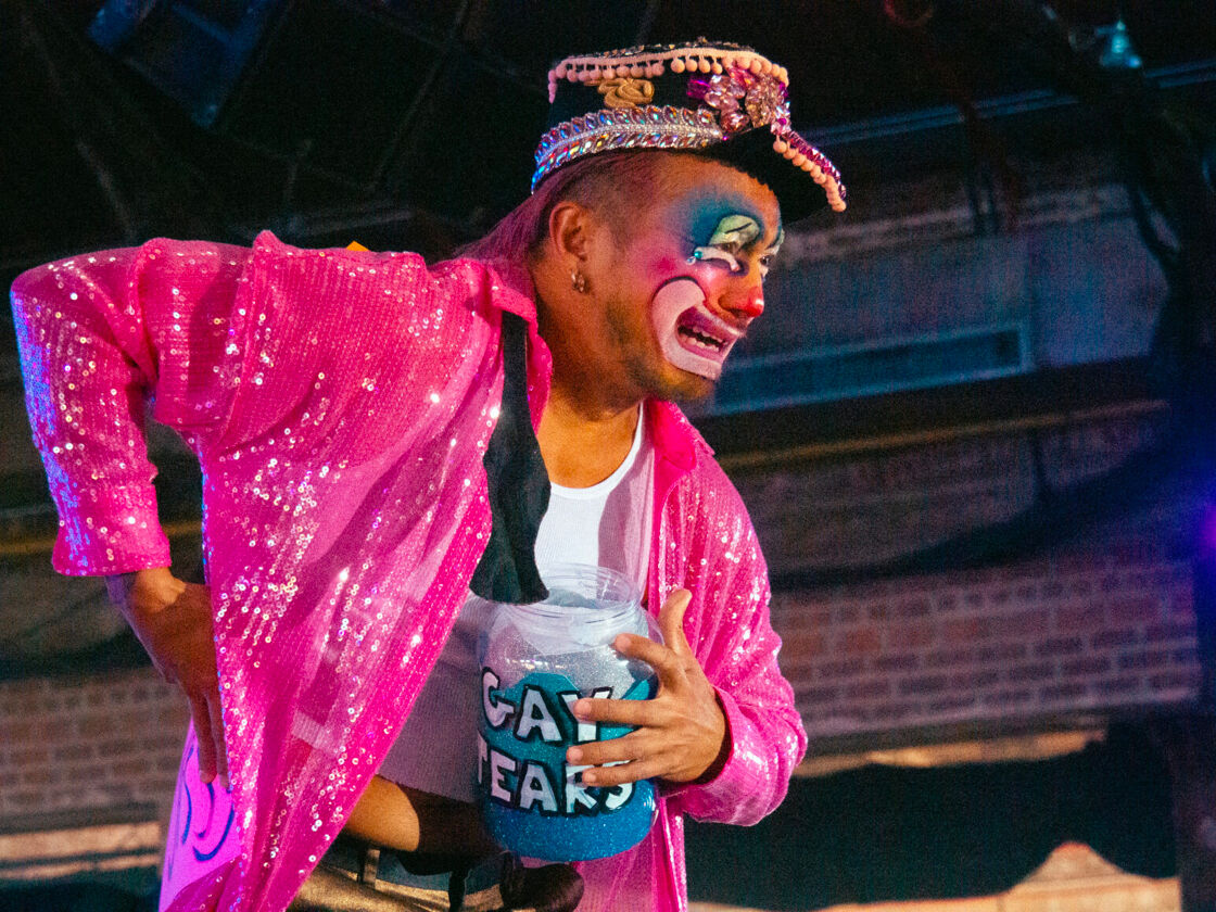 A clown drag queen holds a bucket that says "Gay Tears" at Bushwig. 