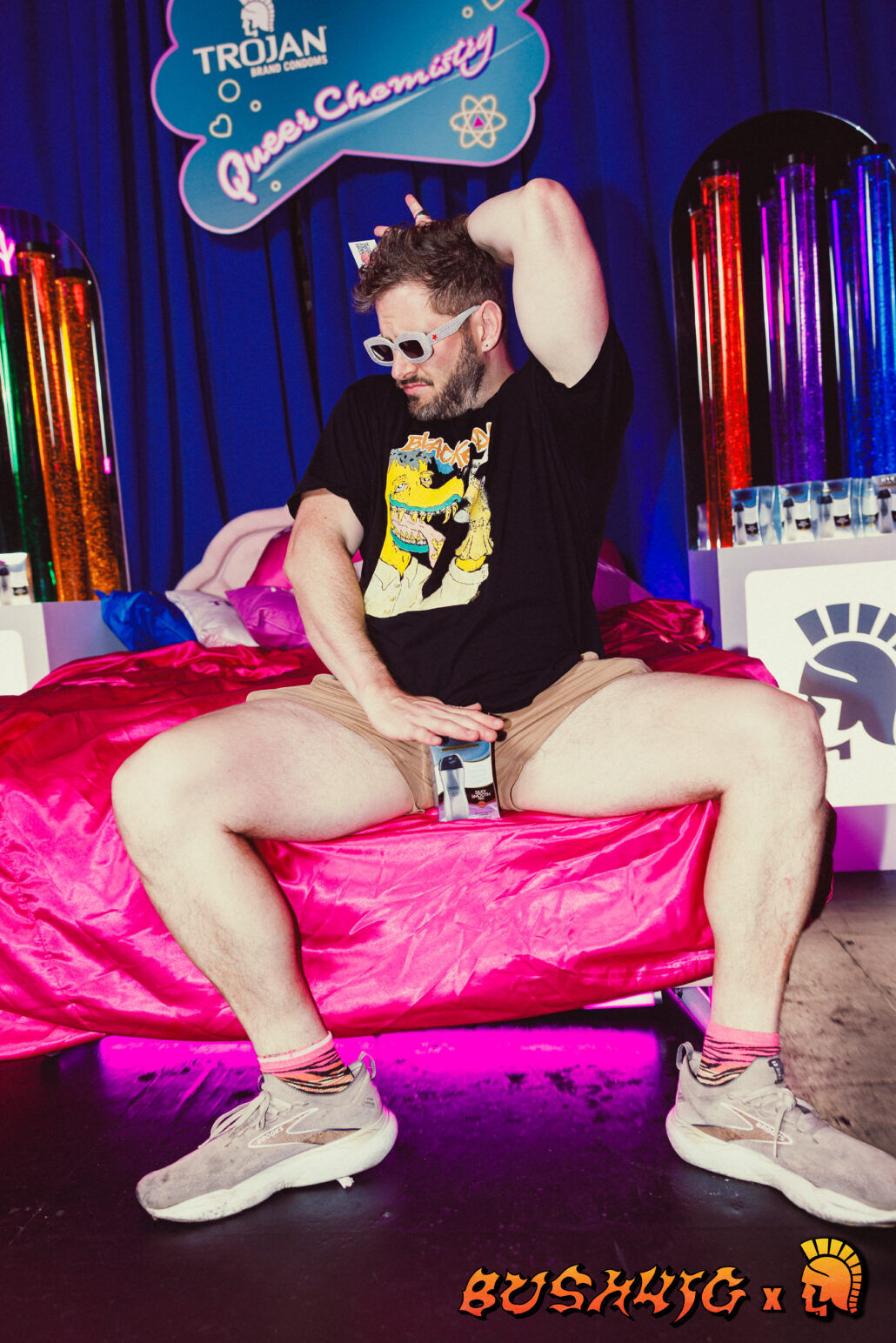 A man poses on a pink comfy bed in front of a Trojan sign at Bushwig.
