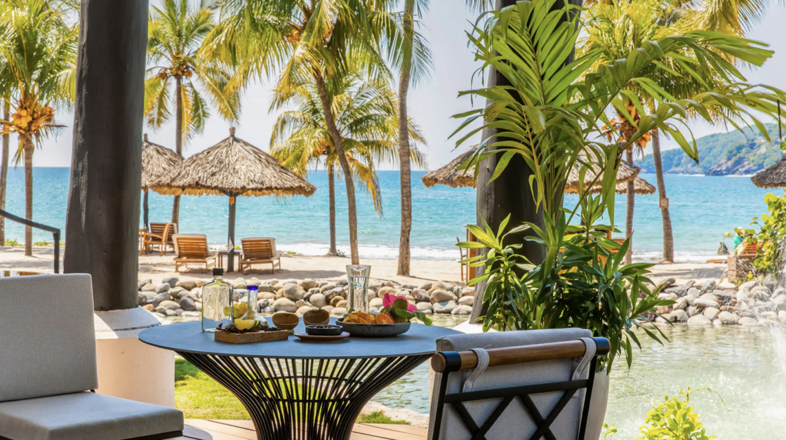 A waterside patio, with table and chairs, looking out at the beach at Thompson Zihuatanejo.