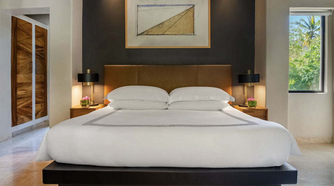 A king bed is pictured inside a room at Thompson Zihuatanejo. On either end, luxe night tables with tabletop lamps on top of them.