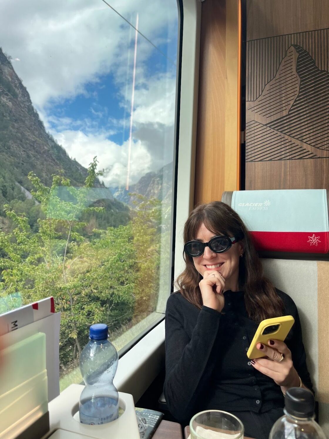 A young woman smiles in a seat of Switzerland's Glacier Express, leaning against a window of scenic views.