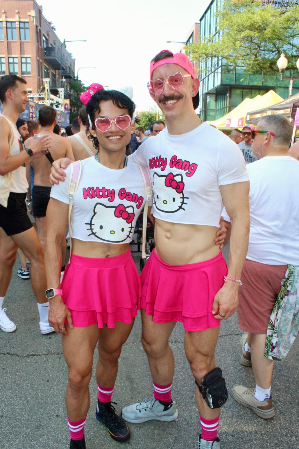 Two men wearing Hello Kitty crop tops that say "Kitty Gang" and short pink skirts smile and pose on the streets of Chicago's Market Days.