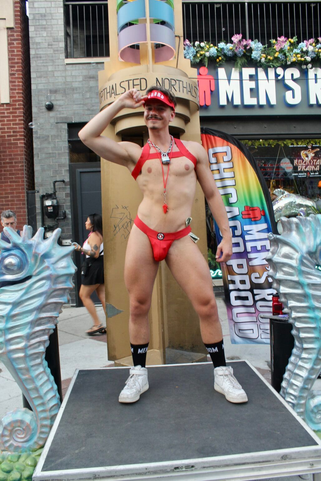 A shirtless man poses in a speedo on a platform, dressed like a lifeguard, at Chicago's Market Days.