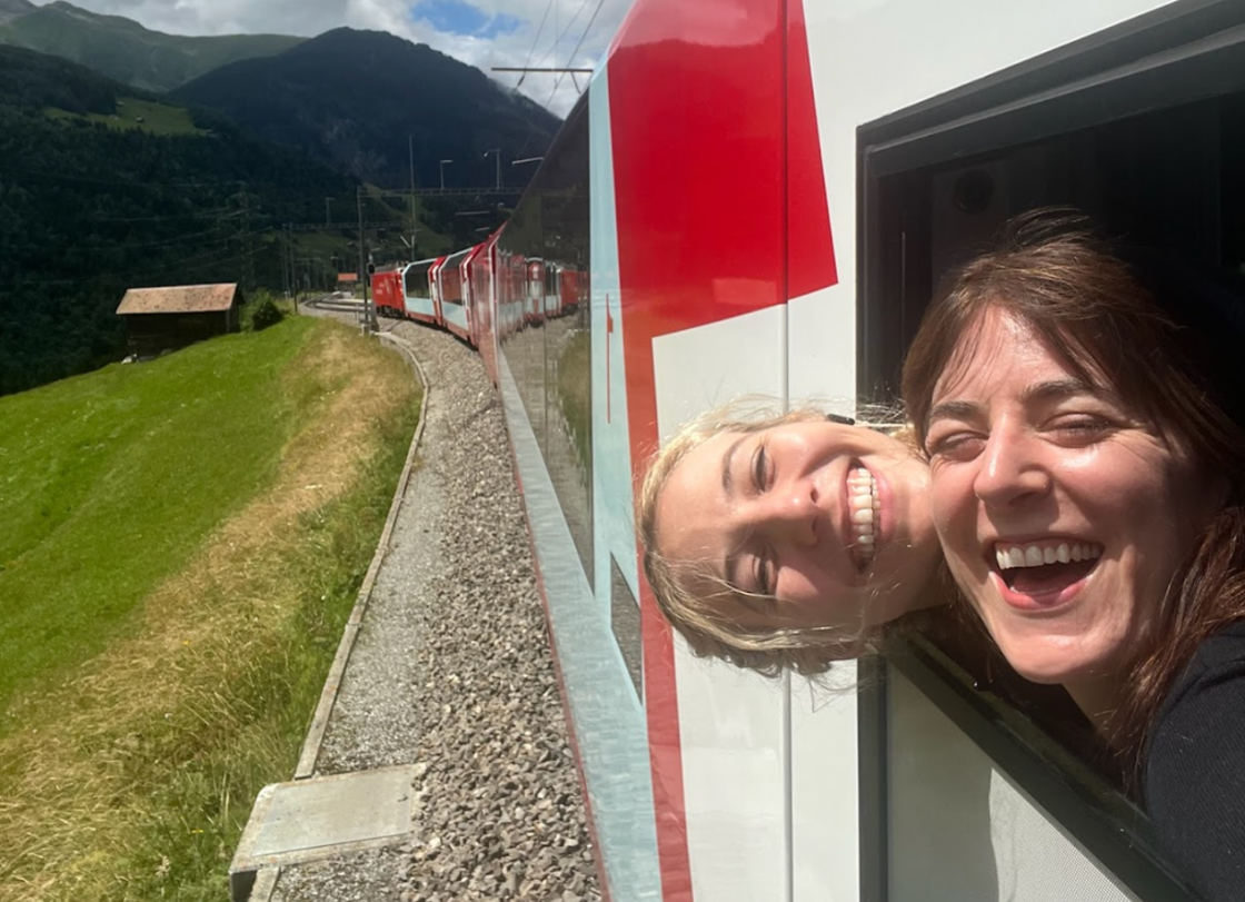 A lesbian couple smiles and sticks their head out of the window of the Glacier Express while riding alongside scenic Switzerland views.