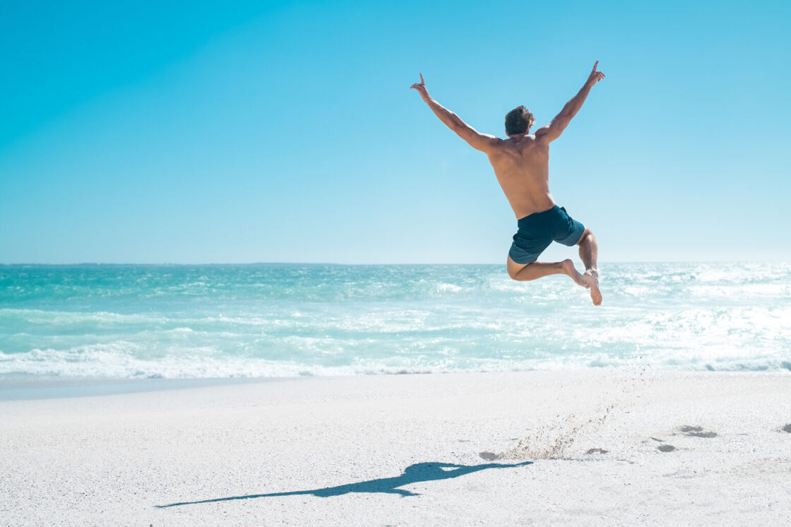 A shirtless man jumps in front of the ocean on a beach.