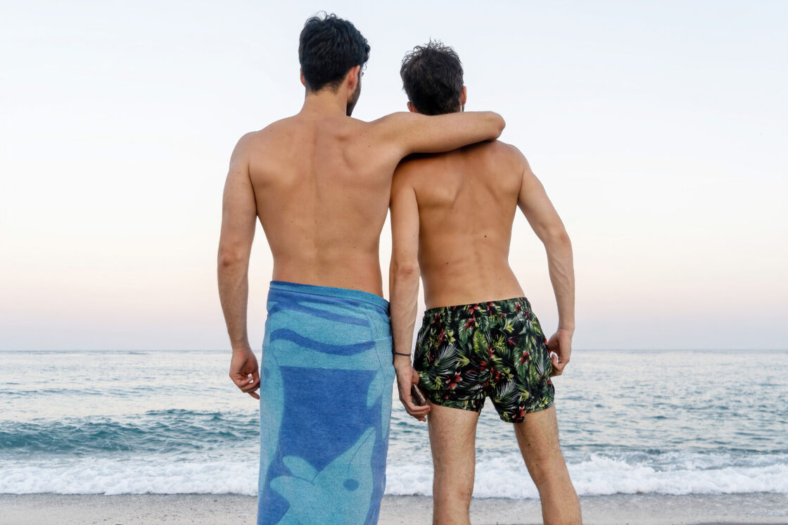 Two shirtless gay men in bathing suits look out toward the ocean while standing on the beach with their arms around each other.