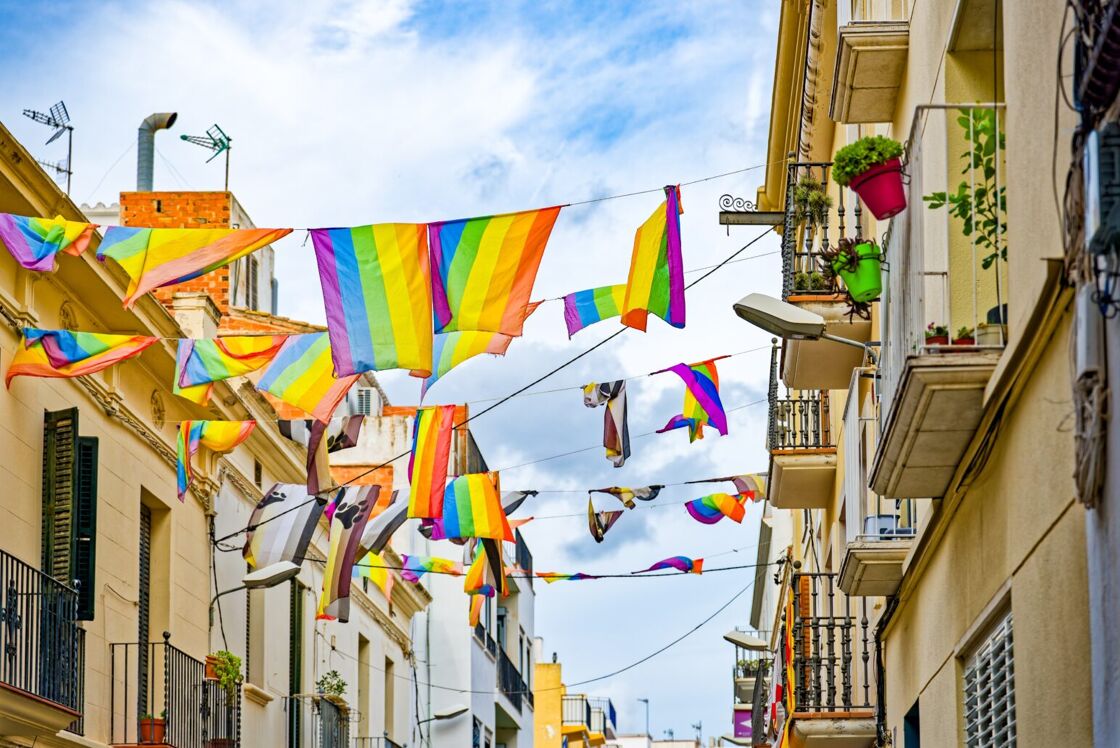 A street in Sitges, Spain, with historic and brightly colored buildings on either side, with Pride flags strewn across the way.