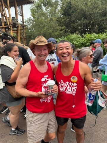 Two gay men smile wearing red tank tops outdoors at Disneyland during Anaheim Gay Days.