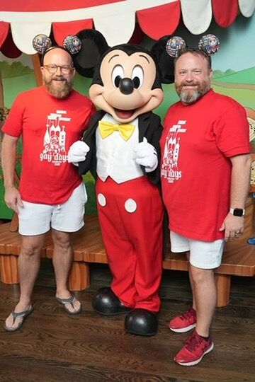 Two gay men pose with Mickey Mouse while wearing red t-shirts at Disneyland during Anaheim Gay Days.