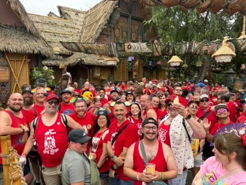 A large group in red poses for a photo in front of the Tiki Room at Disneyland during Anaheim Gay Days.