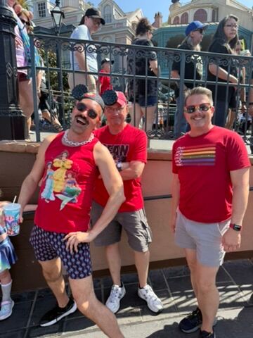 A group of three men smile in red shirts waiting in line at Disneyland during Anaheim Gay Days.