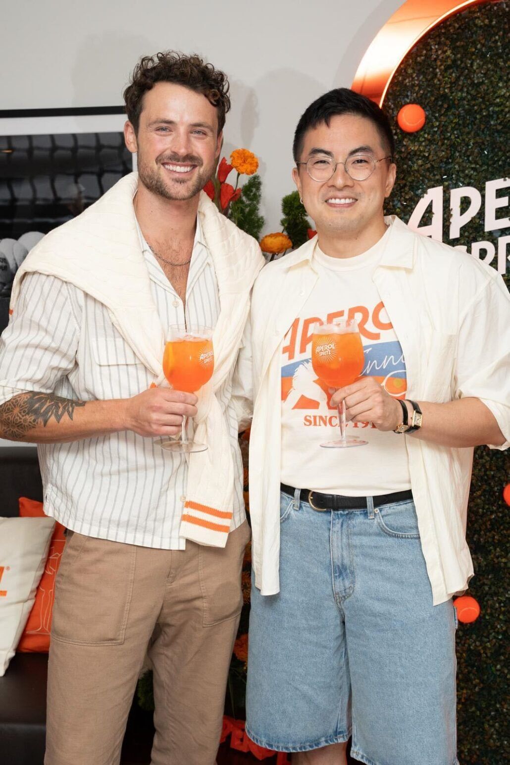 Dylan Efron and Bowen Yang smile holding up Aperol Spritzes at the US Open.