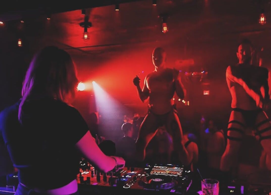 Two male go-go dancers perform on stage at G Lounge in red lighting while a DJ spins tunes in the foreground.