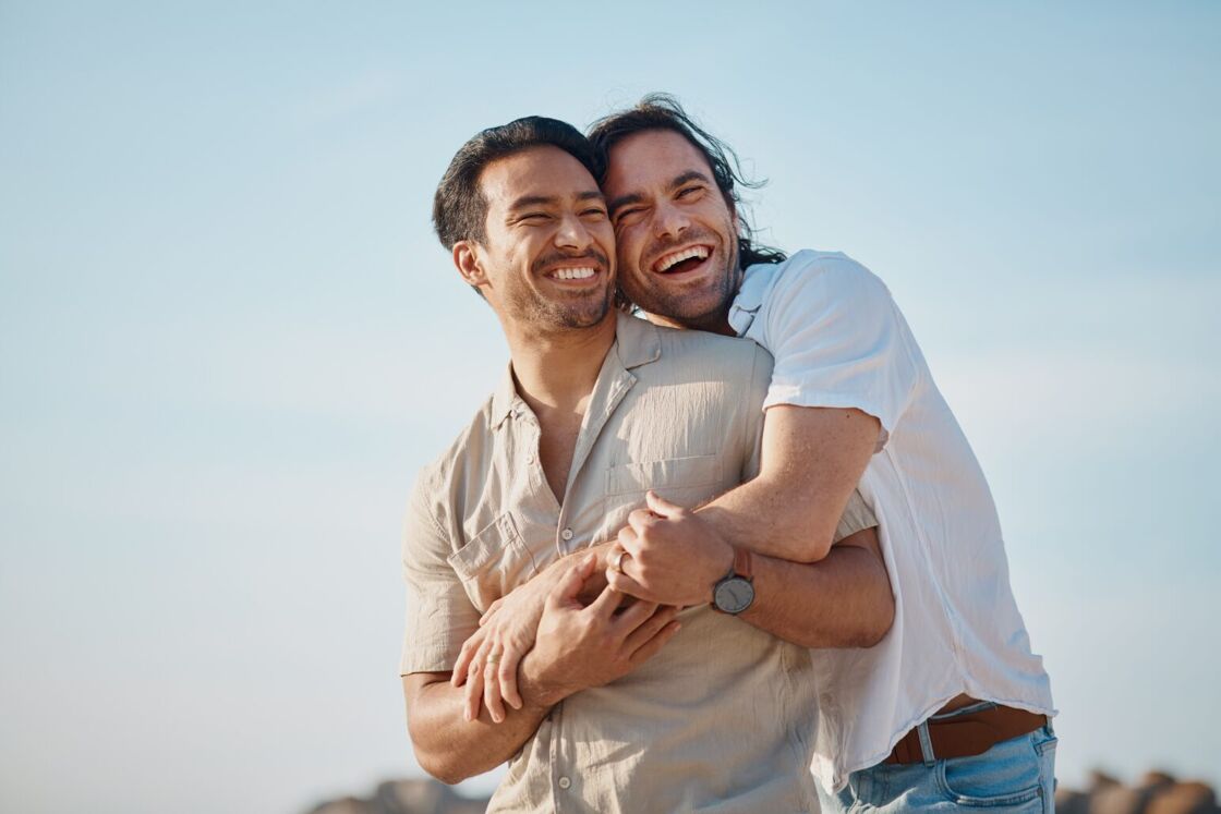 Two gay men embrace on a beach in front of a clear sky.