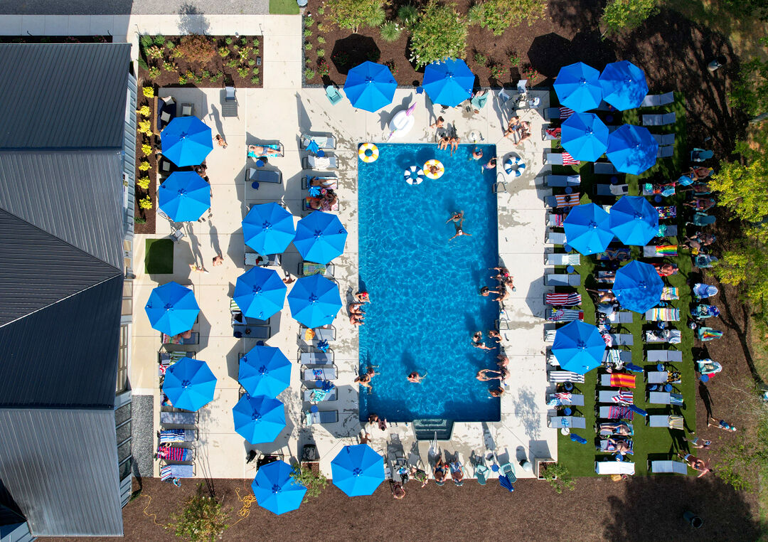 A bird's eye view of Blue Moon Campground's pool and clubhouse, where visitors lounge on chairs and under umbrellas surrounded by green forest.
