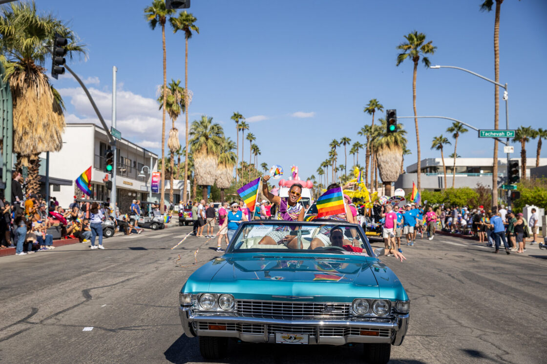 A light blue convertible rolls down the road during Palm Springs Pride, and a young man sticks his head out waving a rainbow flag.