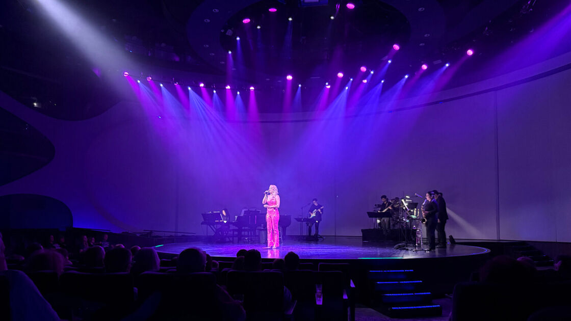 A blonde singer in a pink top and pants performs on stage under blue lights aboard a cruise ship. 
