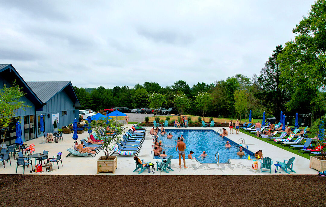 Shirtless men lounge around the pool and clubhouse at Blue Moon Campground, surrounded by lush green forestry.