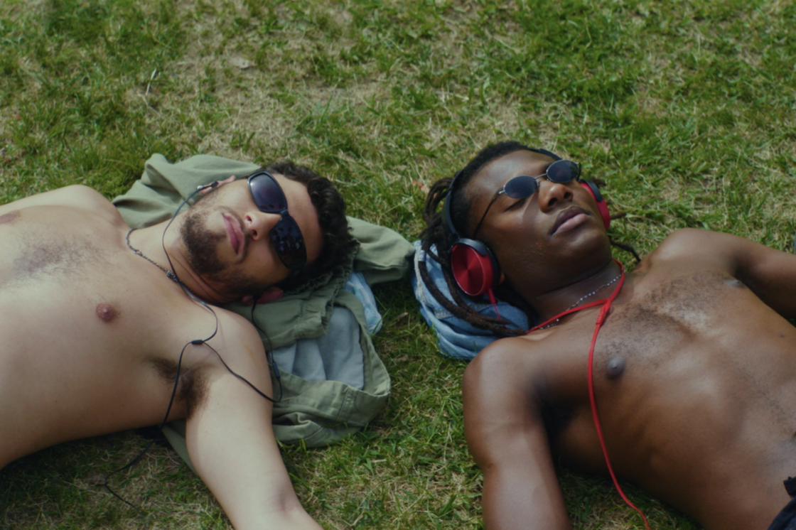 Two shirtless men hold hands on green grass, listening to music on headphones in a scene from "Night Stage."
