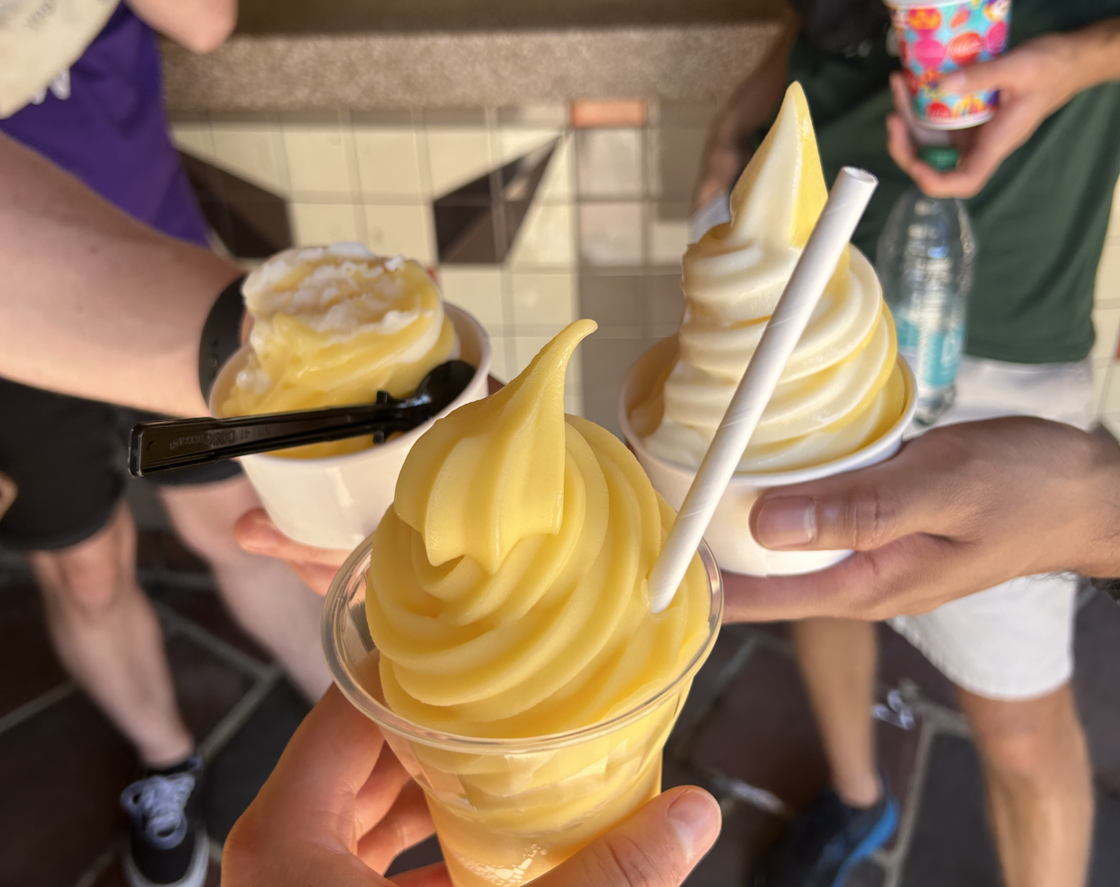 Three men stand in a circle at Disney World showing off their pineapple-flavored Dole Whip soft serve desserts. 