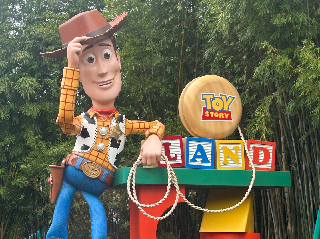 A large statue of Woody from "Toy Story" stands next to a sign reading "Toy Story Land" in front of greenery at Disney's Hollywood Studios.